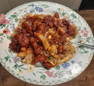 A plate of crispy chicken coated in a thick, glossy red sauce, served over special fried rice. The sauce appears sticky and rich with visible onion pieces, and the rice contains peas and scrambled egg. The dish sits on a floral-patterned plate with a fork on the side.